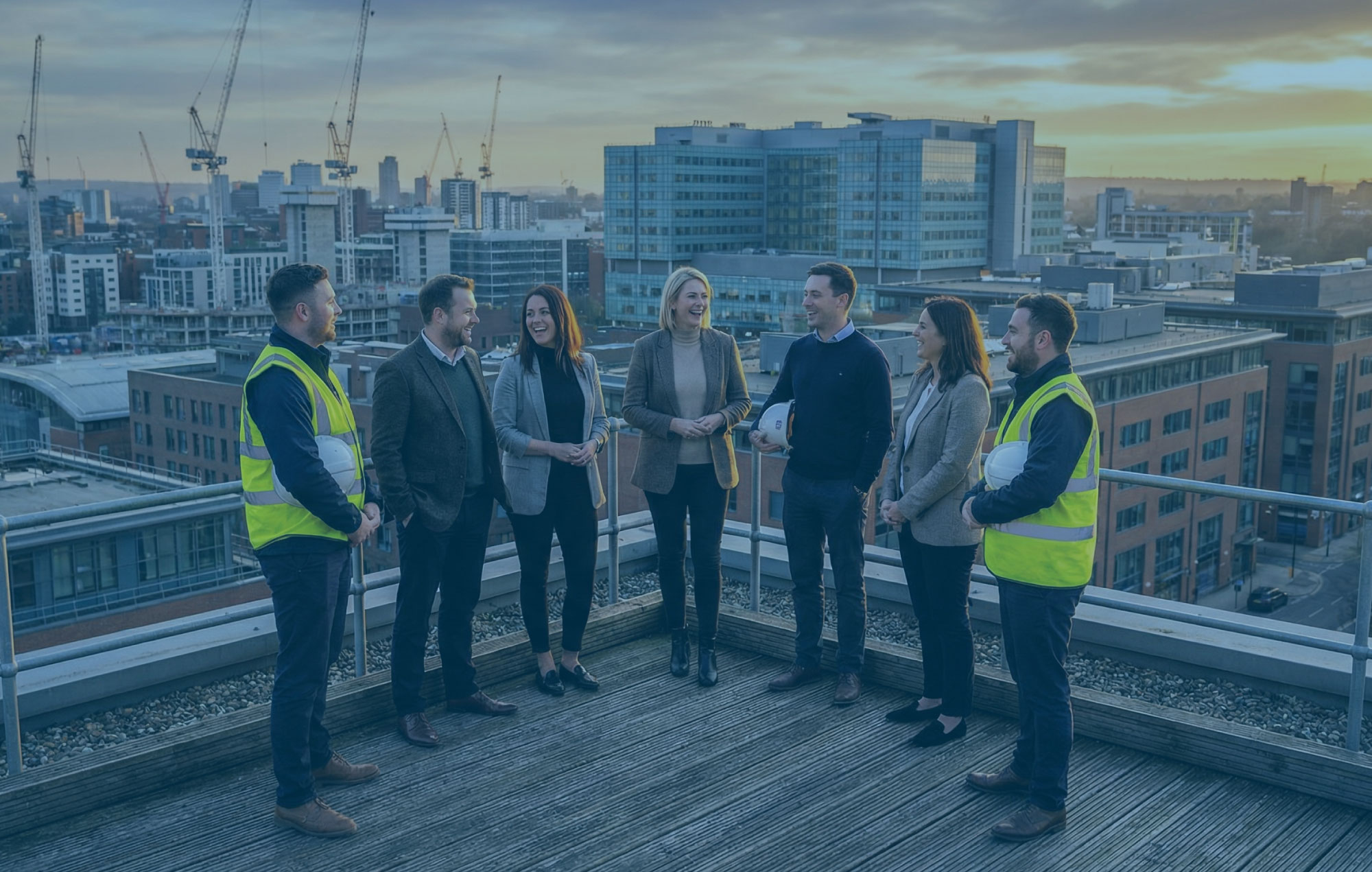 Group of colleagues in PPE and casual wear on a rooftop walkway overlooking a city at sunset, smiling and talking