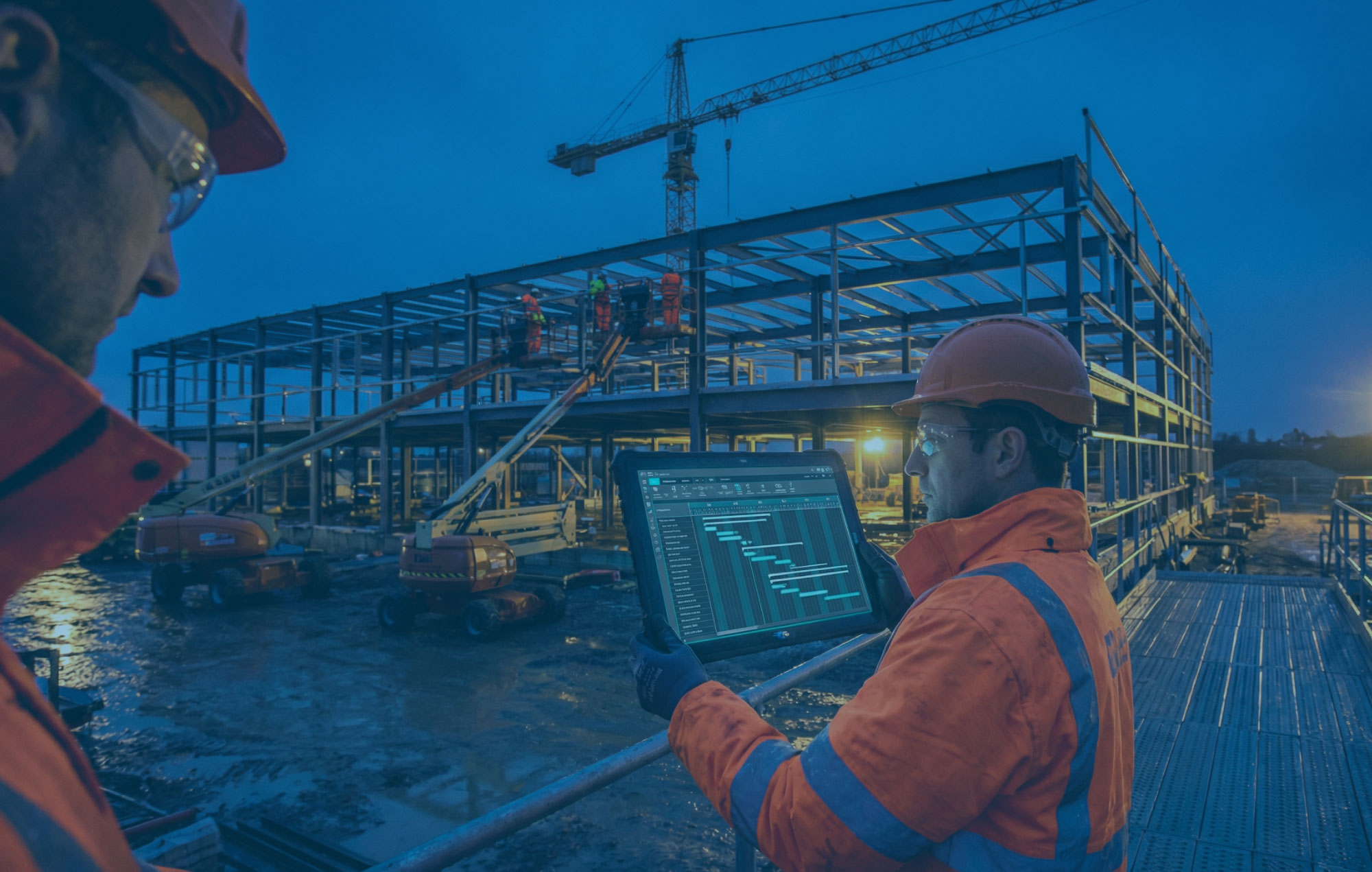 Programme manager on a live construction site checking a tablet schedule with steel frame crane and temporary lighting behind