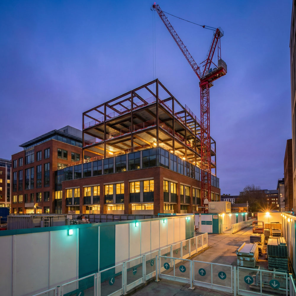 Live construction site at blue hour with steel frame, crane and protected routes around an occupied building